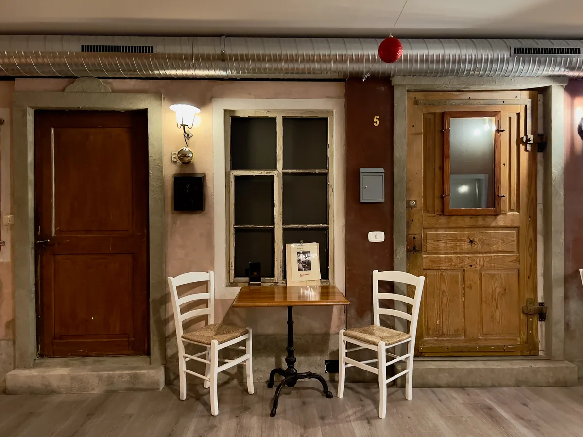 Zwei weisse Holzstühle stehen neben einem quadratischen Tisch mit gusseisernen Füssen vor einer Wand, die wie eine Häuserfassade mit Eingangstüren geschmückt ist.